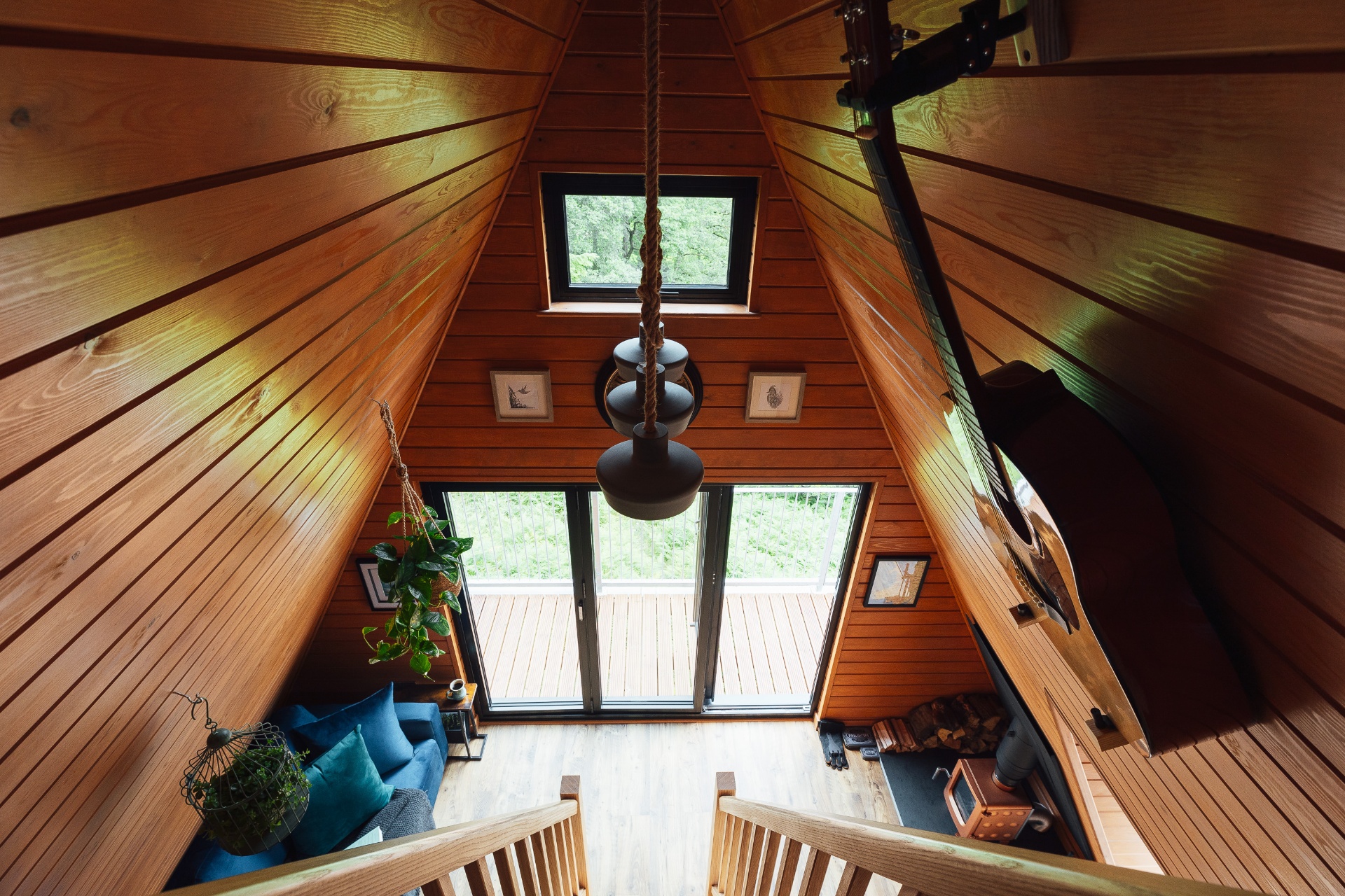 View from the mezzanine looking down through the A-frame rafters to the living space, bifold doors and woodland beyond, with rope-hung pendant lamps in the foreground