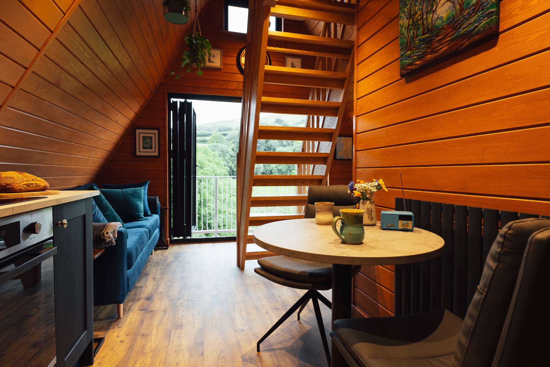 Open-plan view through the cabin from the navy kitchen island past the velvet sofa to the open bifold doors and the staircase rising to the mezzanine, with the green Welsh hills beyond