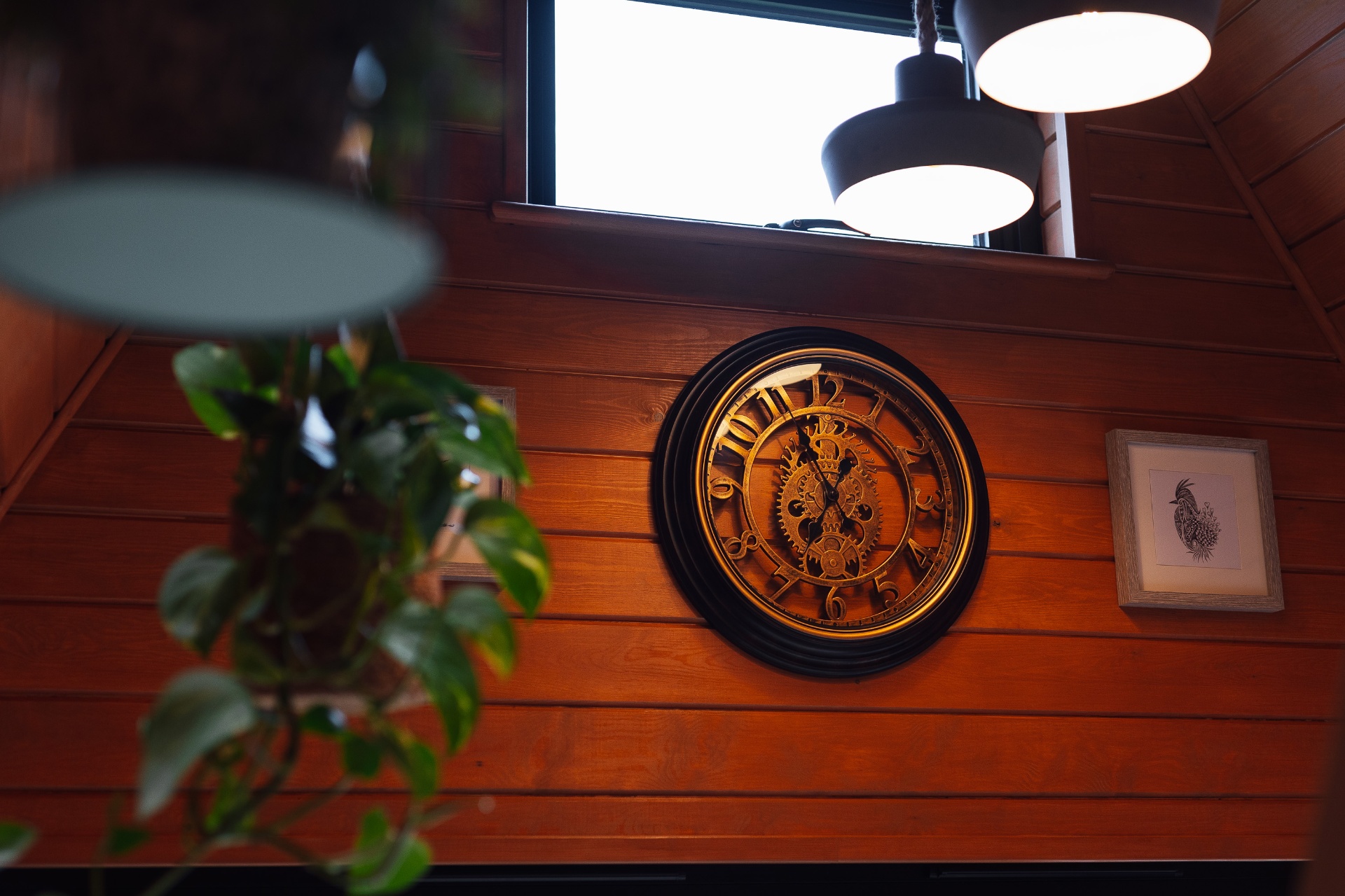 Vintage gold and black wall clock with exposed gears, hung on warm cedar panelling beside a trailing pothos plant and a small framed botanical print inside The Perch