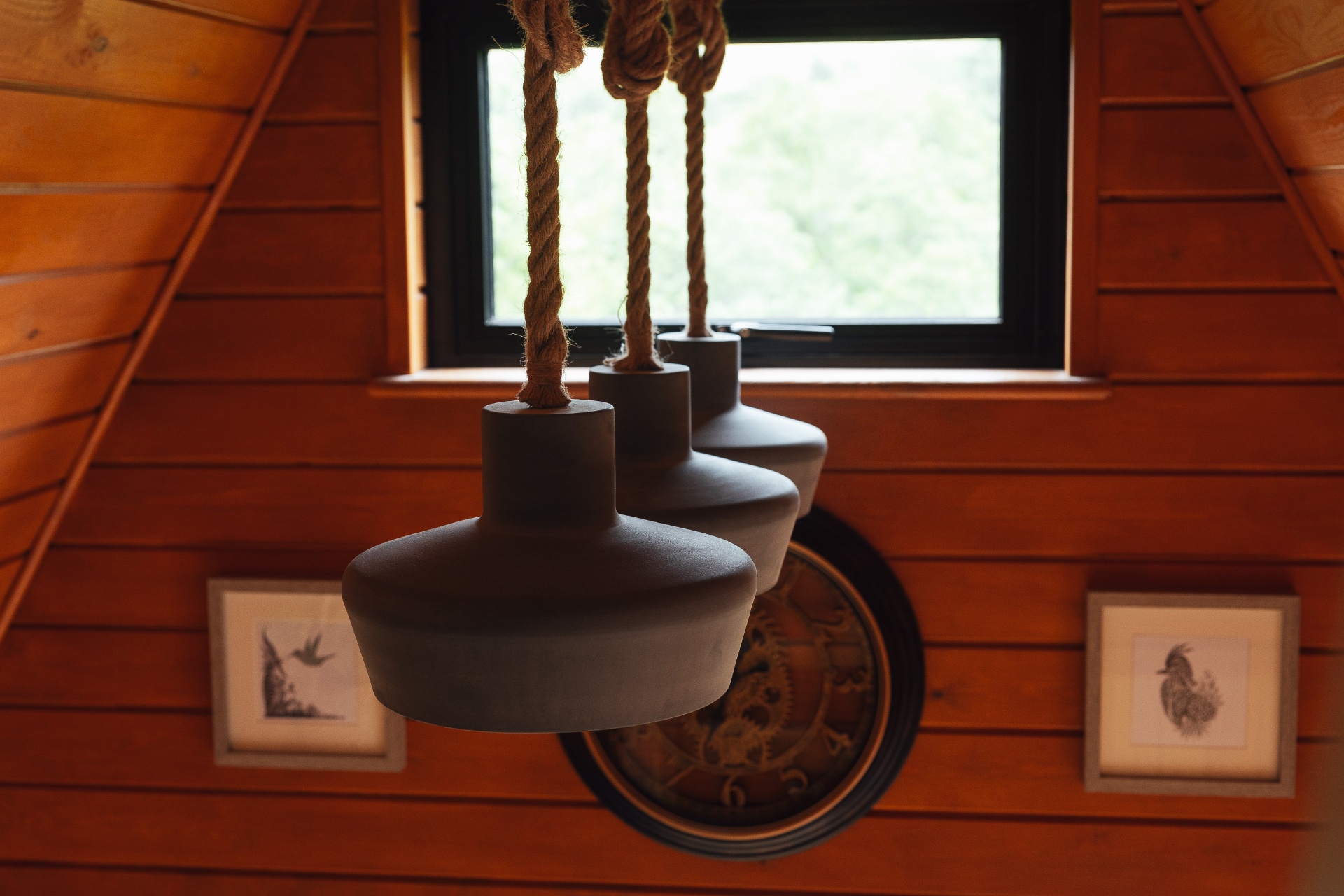 Trio of matte ceramic pendant lamps suspended on rope cords in front of the cedar-clad A-frame wall, with the gear clock and small framed bird prints behind