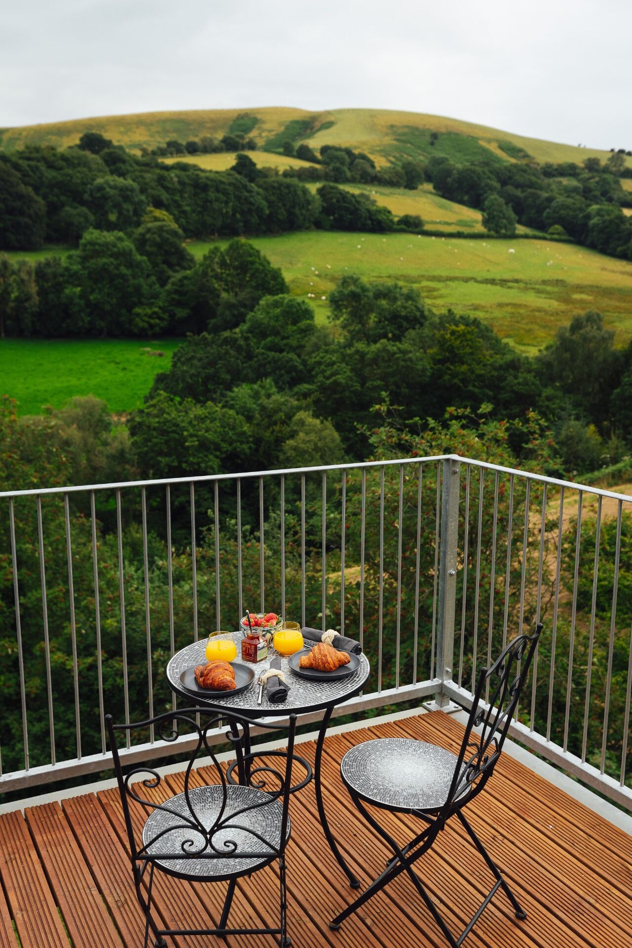Breakfast of croissants, jam and fresh juice on a bistro table on the deck with rolling Welsh hills beyond