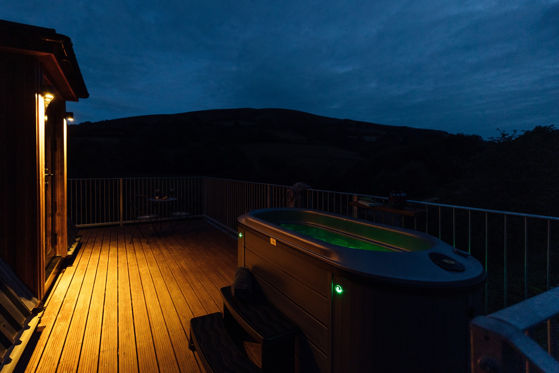 Private hot tub glowing green on the lit cabin deck at dusk, with a bistro table for two and the silhouette of the Welsh hills under a deep blue sky
