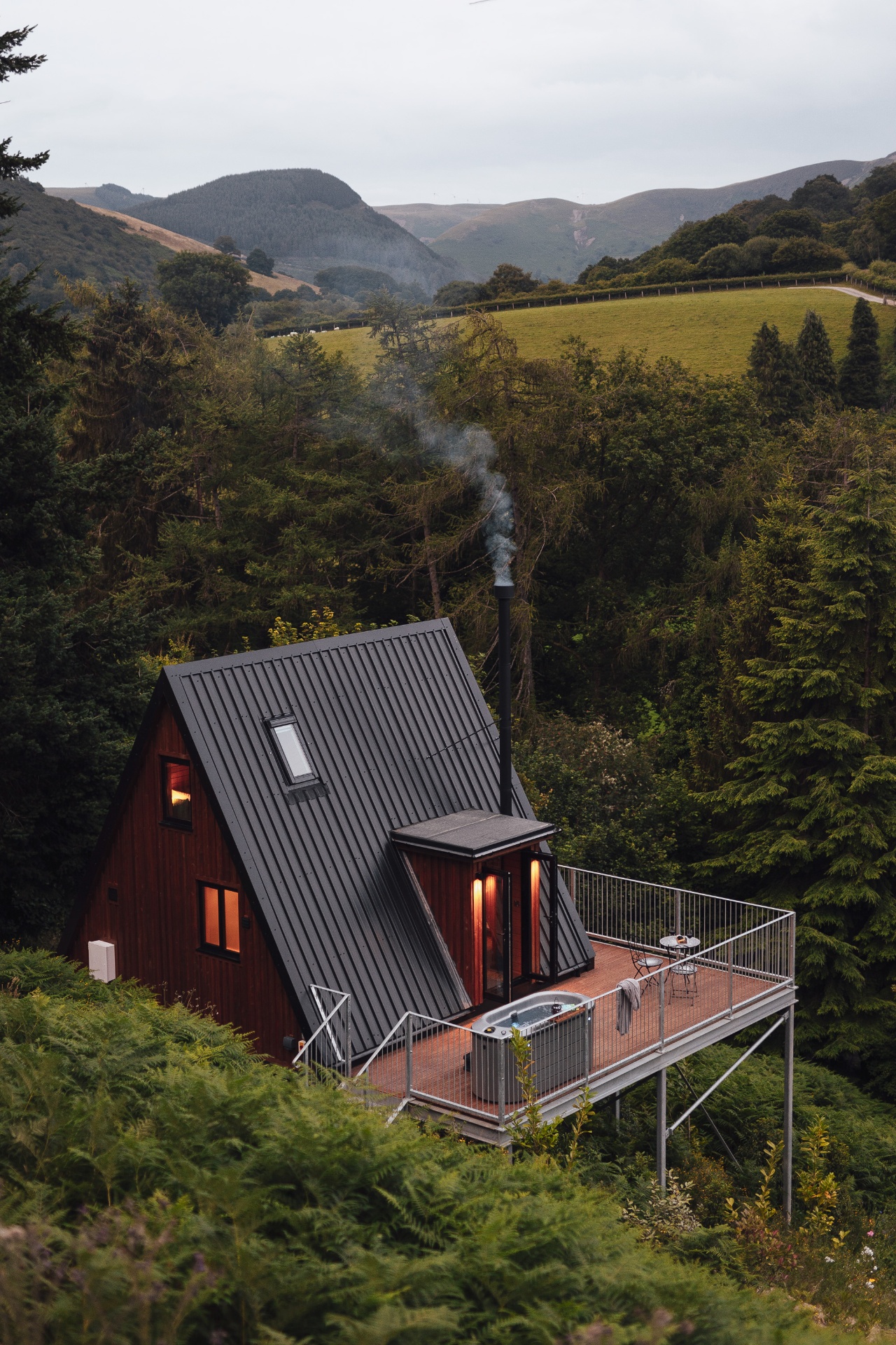 Aerial view of the dark A-frame cabin with smoking wood burner chimney and hot tub on the deck, nestled in woodland above a green Welsh valley with rolling hills beyond